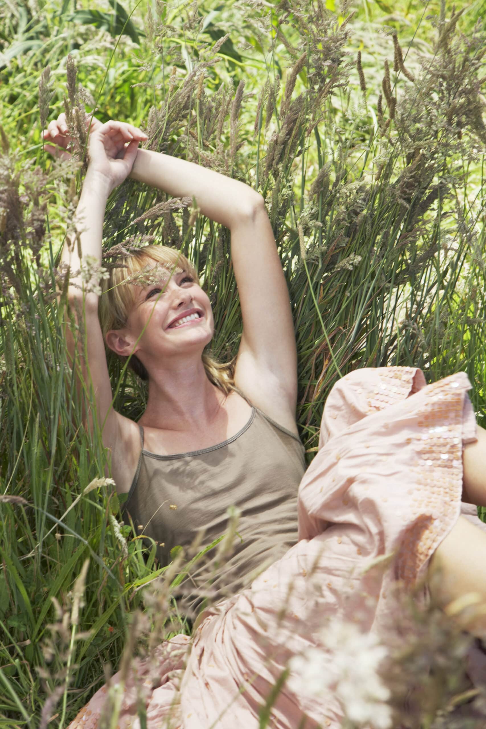 Eine fröhliche Frau in einem leichten Kleid liegt im hohen Gras, lächelt und streckt die Arme über den Kopf, umgeben von üppigem Grün. © Fotografie Tomas Rodriguez