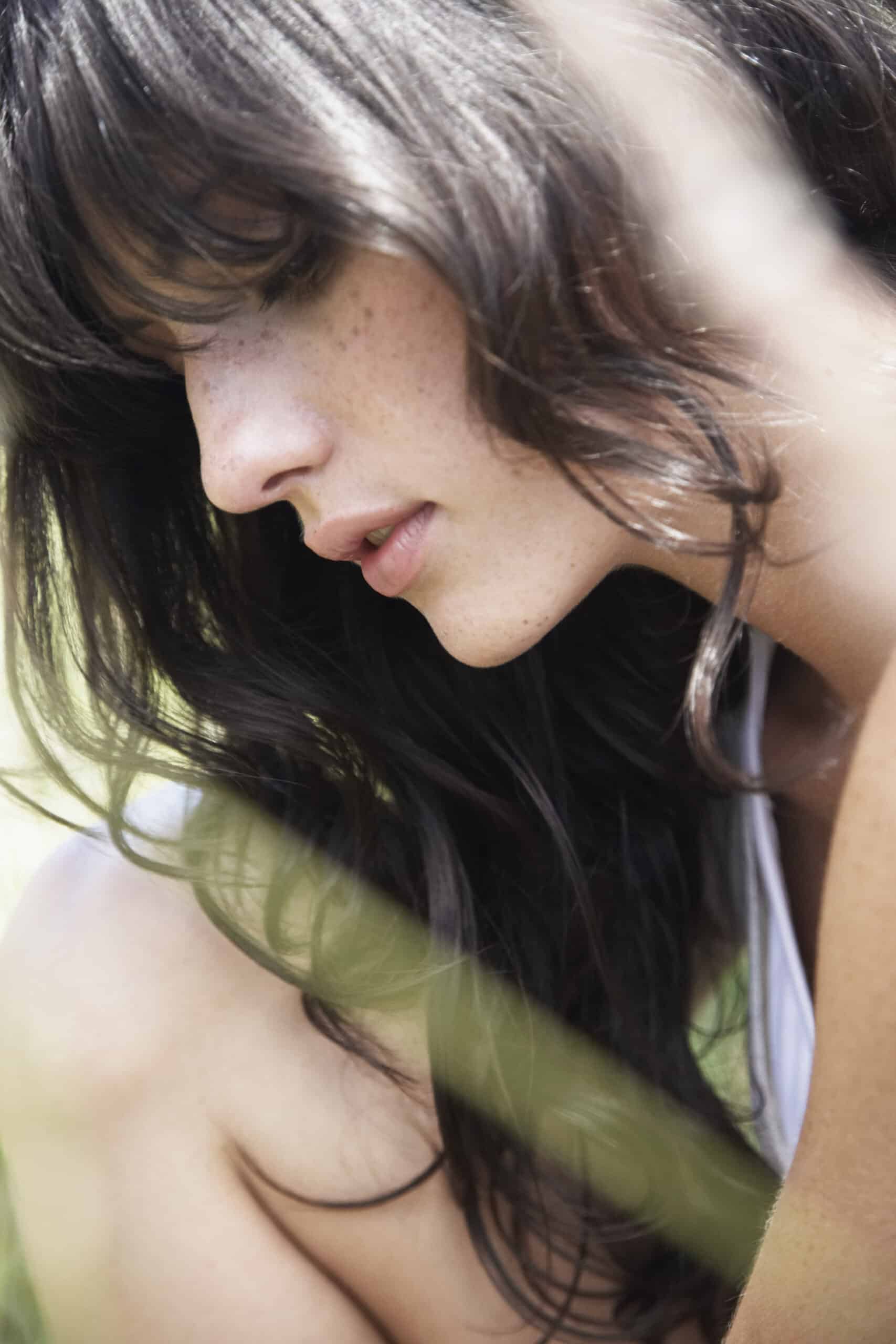 Ein Nahaufnahmeporträt einer Frau mit Sommersprossen, deren Blick nach unten gerichtet ist und teilweise durch sanftes Grün im Vordergrund verdeckt wird. © Fotografie Tomas Rodriguez