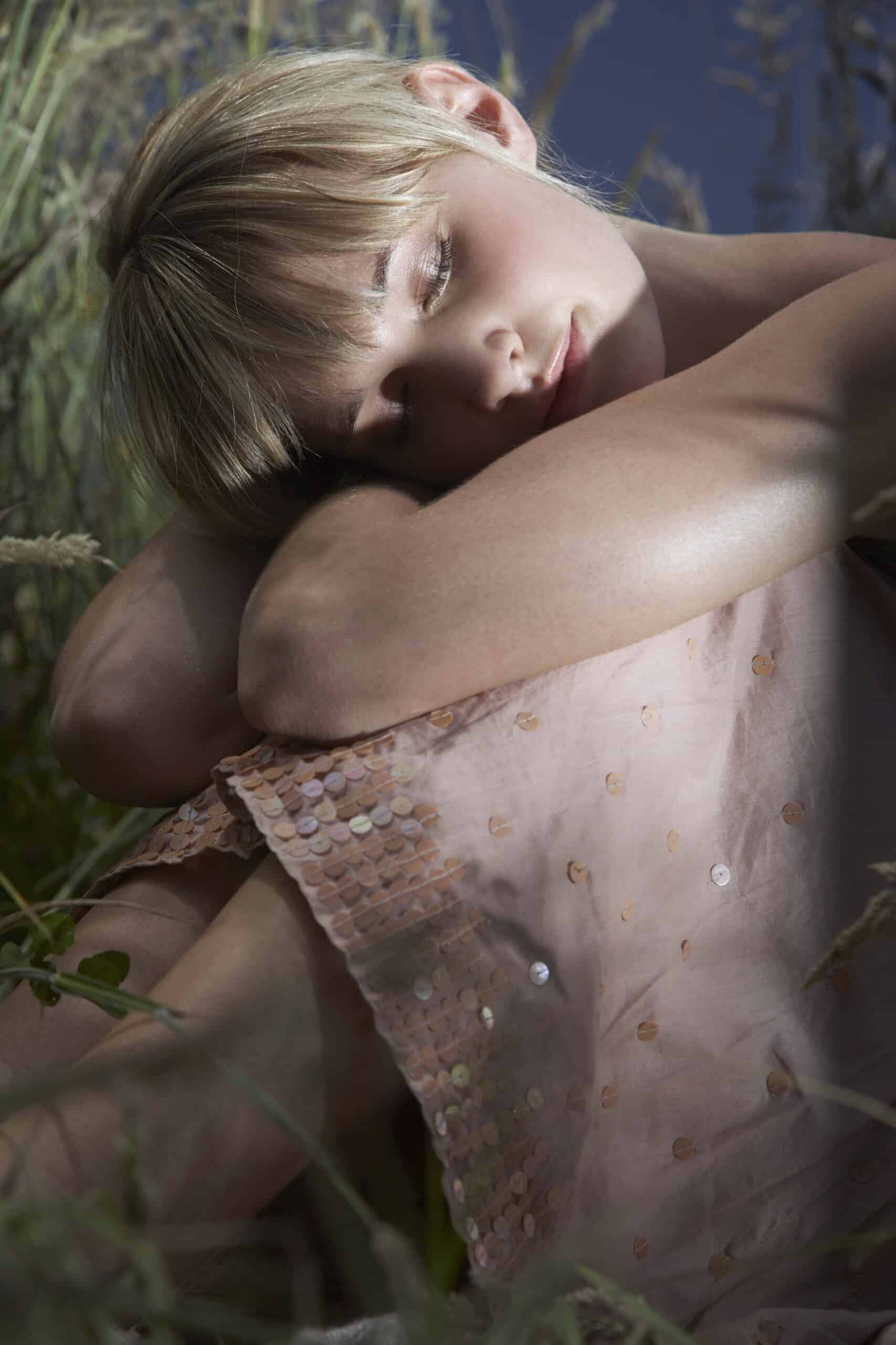 Eine junge Frau mit kurzen blonden Haaren ruht inmitten von hohem Gras und Wildblumen auf ihren Armen und trägt ein zartrosa, mit Pailletten verziertes Kleid. © Fotografie Tomas Rodriguez