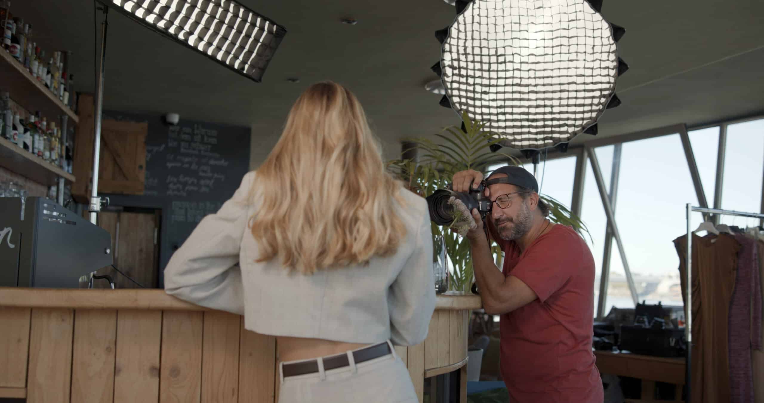 Ein Fotograf mit rotem Hemd und Brille fotografiert ein weibliches Model mit langen blonden Haaren und grauem Anzug. Sie befinden sich in einem Studio mit professioneller Beleuchtungsausrüstung und verschiedenen Requisiten um sie herum. © Fotografie Tomas Rodriguez