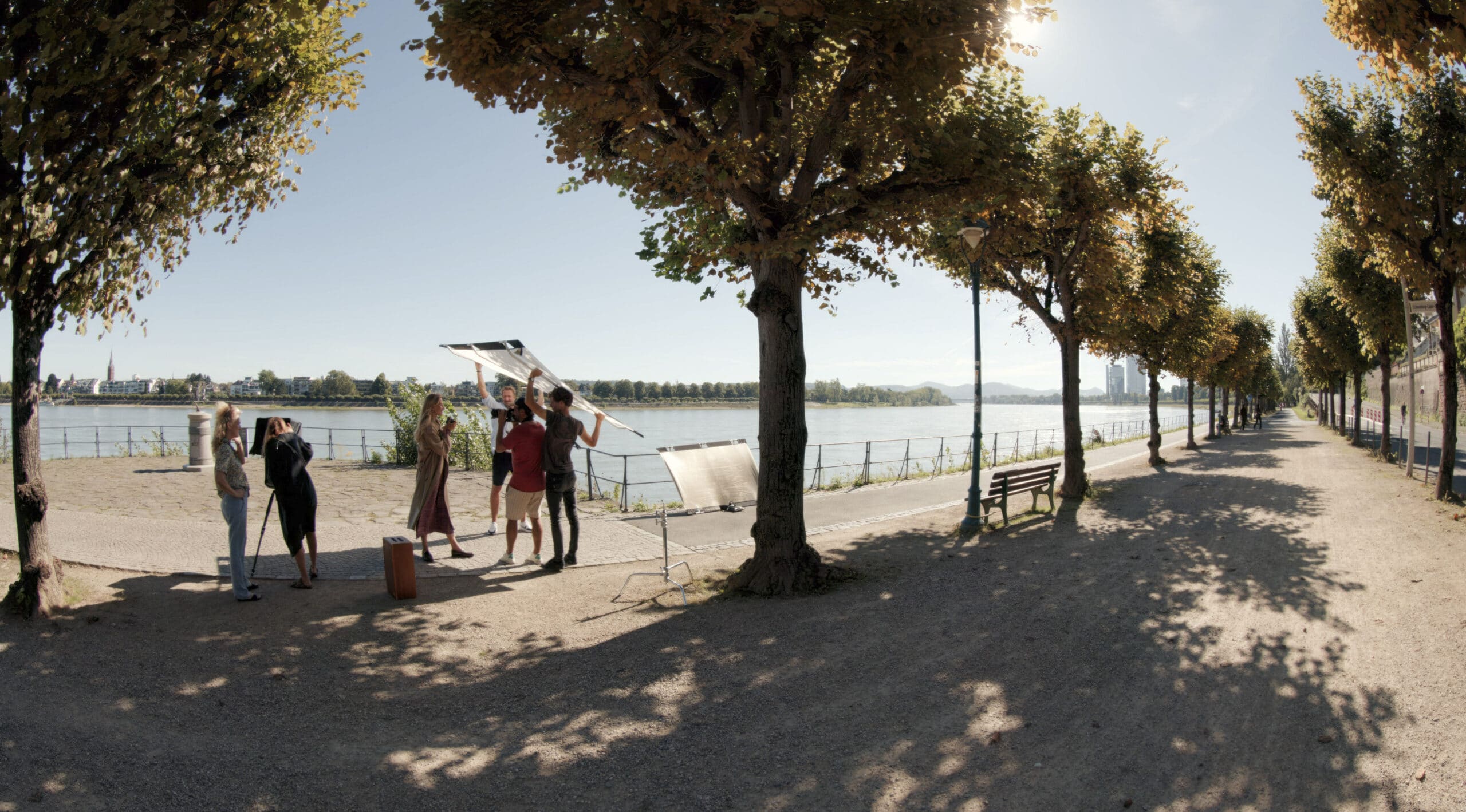 Fünf Personen versammelten sich unter Bäumen an einem sonnenbeschienenen Flussuferweg und arbeiteten oder diskutierten über etwas, das mit reflektierenden Platten zu tun hatte. Im Hintergrund sind ein Geländer am Fluss, entfernte Gebäude und Berge unter einem klaren Himmel zu sehen. © Fotografie Tomas Rodriguez