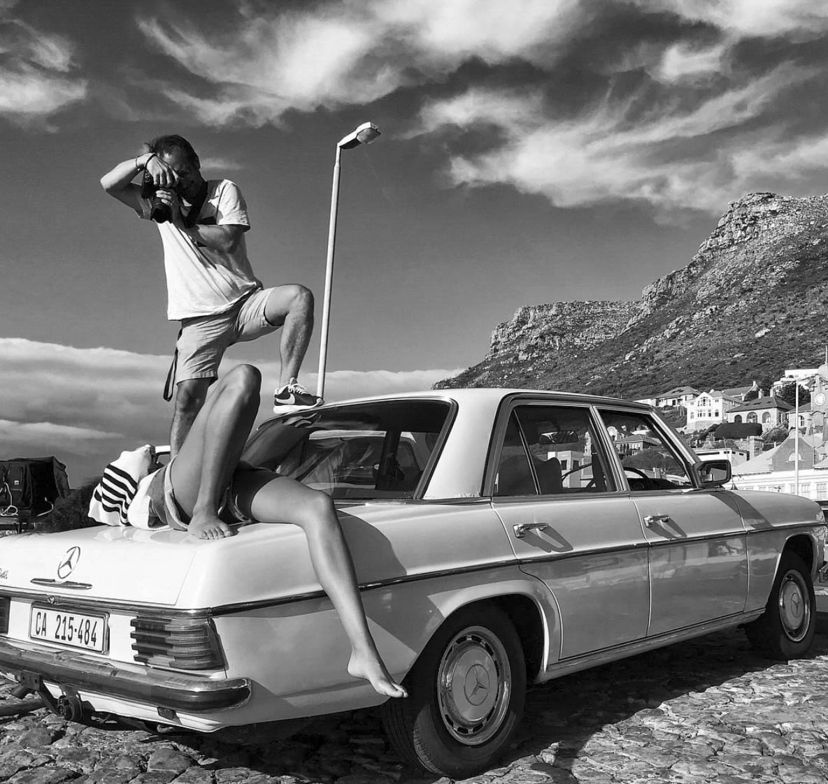 Ein Schwarzweißfoto eines Mannes, der auf einem alten Mercedes steht und mit einer Kamera Fotos macht. Eine Frau liegt auf dem Kofferraum des Autos. Im Hintergrund sind Berge, ein bewölkter Himmel und ein paar Gebäude zu sehen. Auf dem Nummernschild steht „CA 215-464“. © Fotografie Tomas Rodriguez