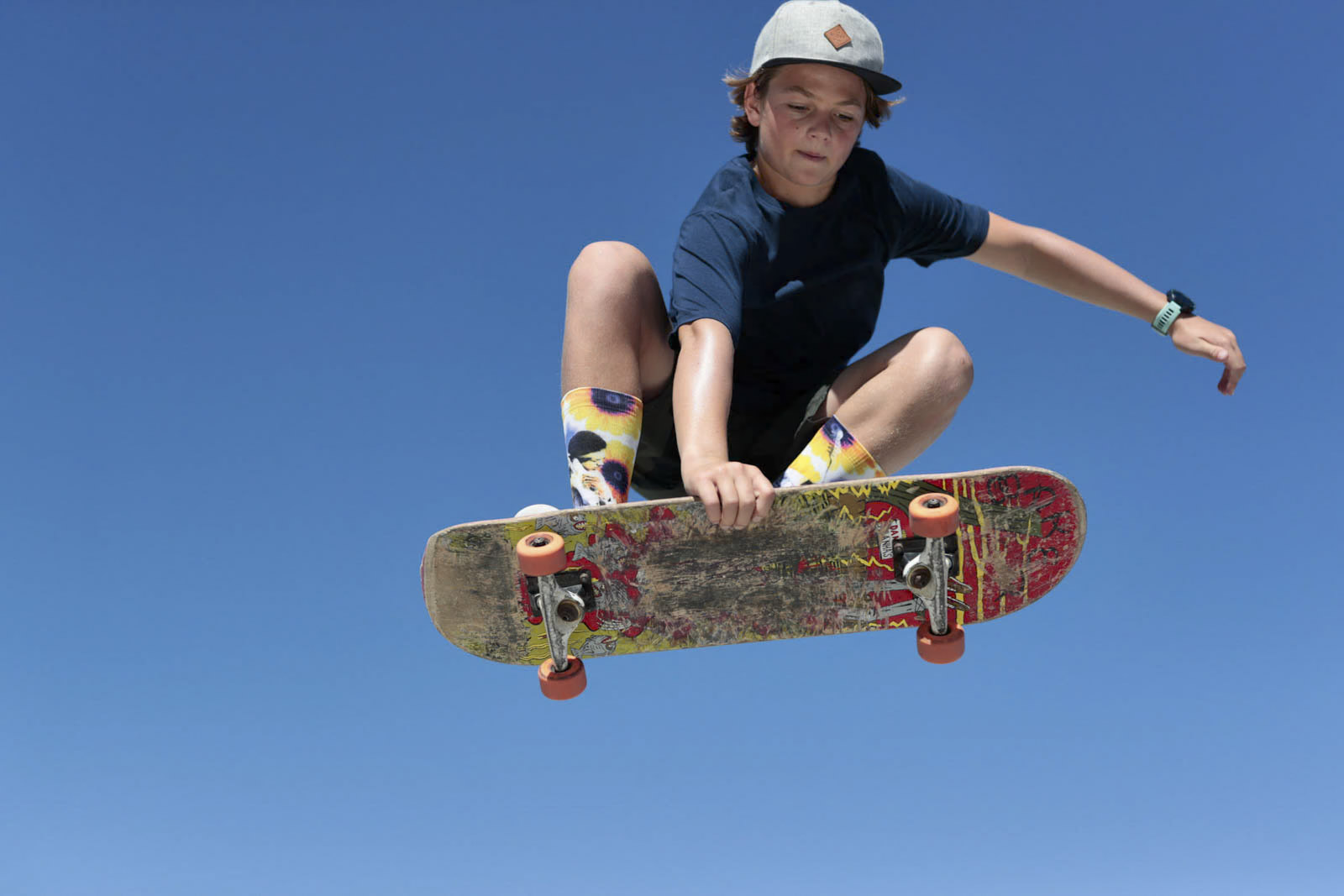 Eine Person mit einer grauen Kappe führt vor einem klaren blauen Himmel einen Trick in der Luft auf einem Skateboard aus. Der Skateboarder trägt ein dunkelblaues Hemd, bunt gemusterte Socken und eine Armbanduhr und stellt damit sein Können und sein Gleichgewicht zur Schau. © Fotografie Tomas Rodriguez