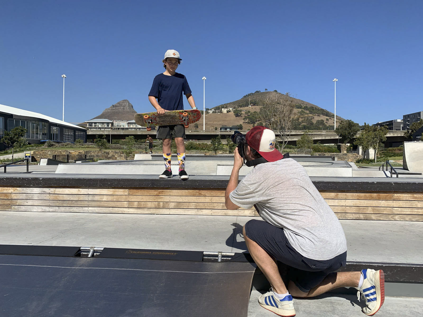 Eine Person mit Hut und bunten Socken steht mit einem Skateboard auf einem Felsvorsprung, während eine andere Person auf dem Boden kniet und ein Foto von ihnen in einem Skatepark macht. Im Hintergrund sind ein klarer blauer Himmel und ein Hügel zu sehen.
. © Fotografie Tomas Rodriguez