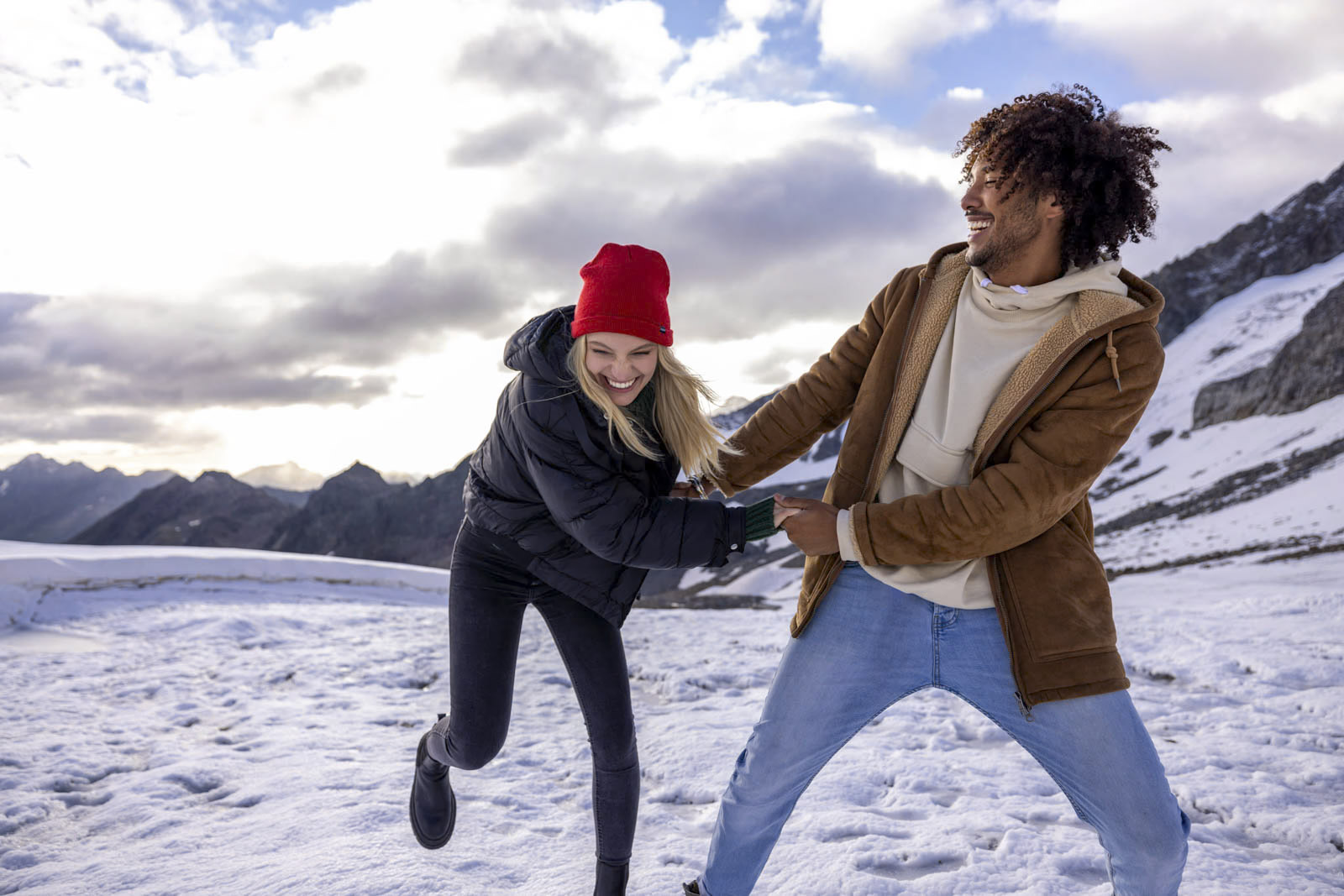 Eine Frau in einer schwarzen Jacke und einer roten Mütze und ein Mann in einer braunen Jacke und Jeans halten verspielt Händchen und lachen in einer schneebedeckten Bergregion mit einem bewölkten Himmel im Hintergrund. © Fotografie Tomas Rodriguez