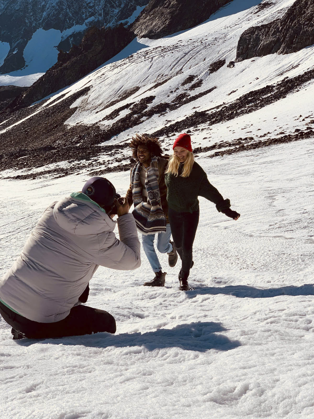 Zwei Frauen in Winterkleidung laufen zusammen und lachend einen verschneiten Berghang entlang. Die eine trägt einen gemusterten Mantel, die andere eine rote Mütze. Eine kniende Person in einer weißen Jacke macht ein Foto von ihnen. Im Hintergrund sind felsige Gipfel zu sehen. © Fotografie Tomas Rodriguez