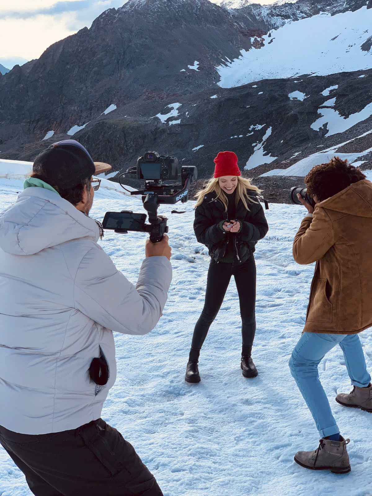 Ein Model steht auf einem schneebedeckten Berg und trägt eine rote Mütze und eine schwarze Jacke. Zwei Fotografen fokussieren auf sie, einer hält eine Kamera auf einem Stabilisator und ein anderer eine Handkamera. Felsige Berge und Schneeflecken bilden den Hintergrund. © Fotografie Tomas Rodriguez