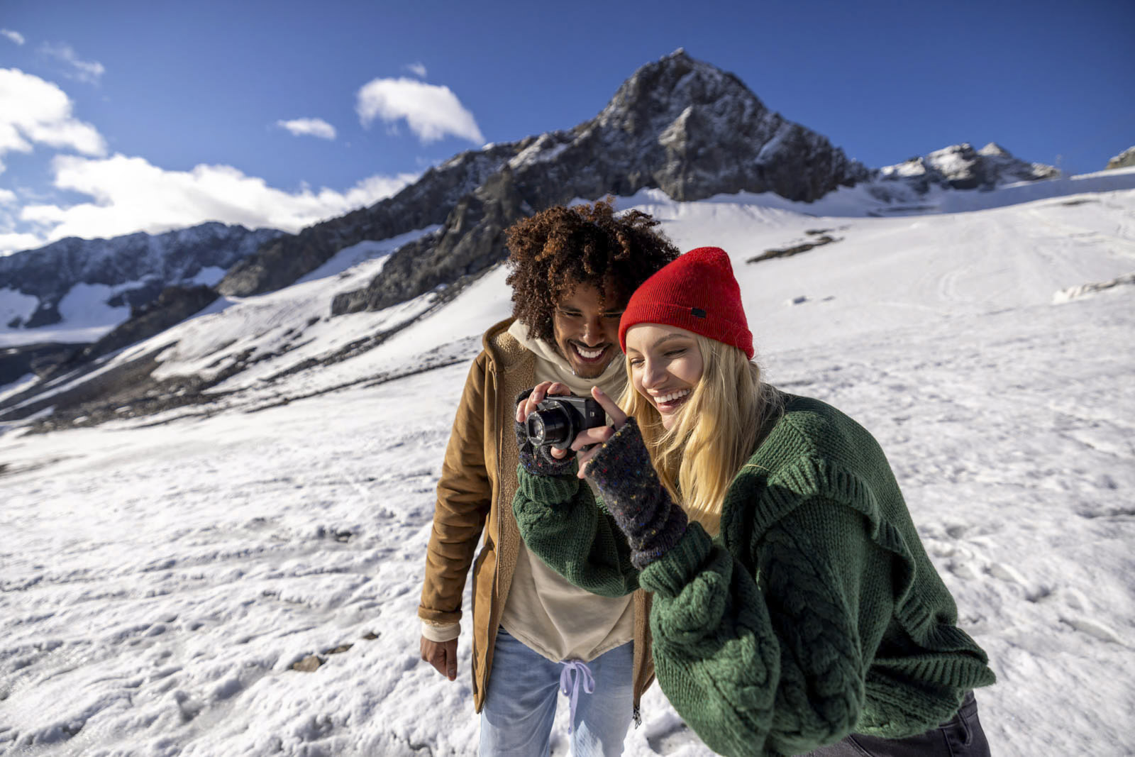 Ein lächelndes Paar steht auf einem schneebedeckten Berg. Die Frau mit der roten Mütze und dem grünen Pullover zeigt dem Mann in der braunen Jacke etwas auf einer Kamera. Der Himmel ist klar und die Berggipfel sind mit Schnee bedeckt. © Fotografie Tomas Rodriguez