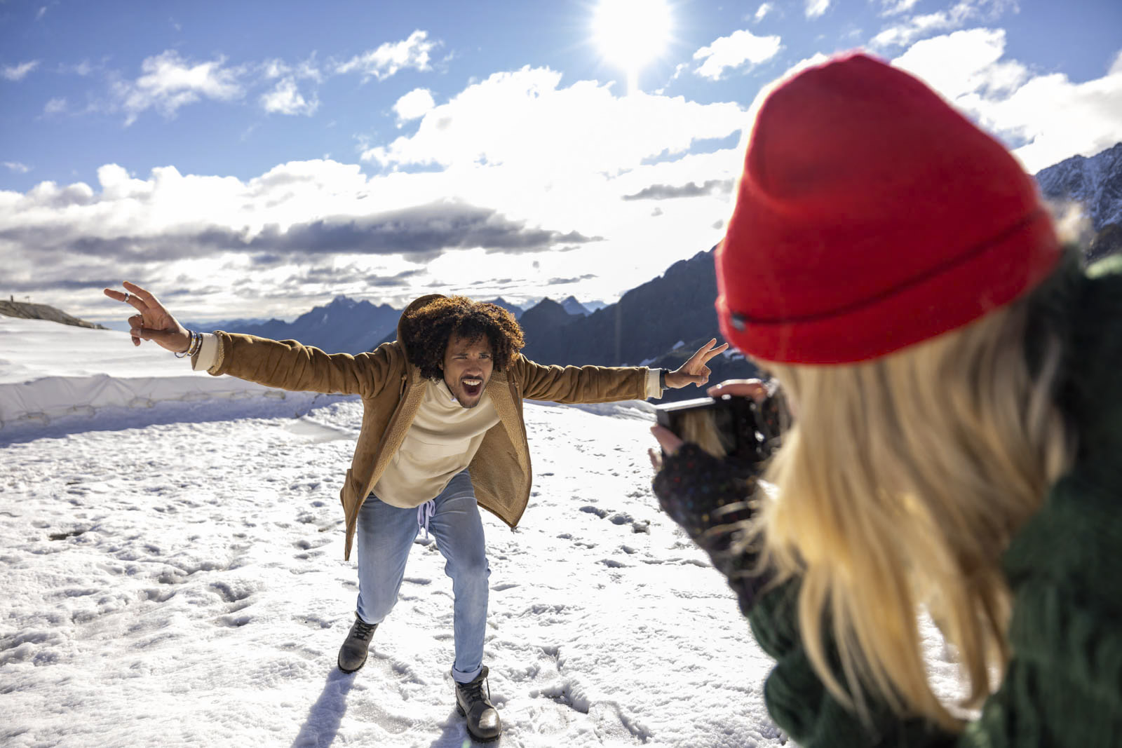 Eine Person in einem braunen Mantel und einem weißen Pullover posiert verspielt auf einem schneebedeckten Berg und streckt die Arme weit aus. Eine andere Person in einer roten Mütze und einer dunkelgrünen Jacke macht ein Foto von ihr. Der Himmel ist hell und teilweise bewölkt, im Hintergrund sind Berge zu sehen. © Fotografie Tomas Rodriguez