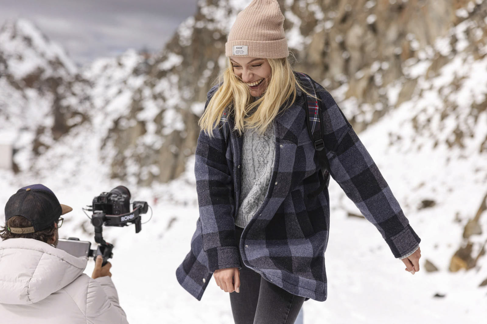 Eine Frau mit Beanie und kariertem Mantel lächelt, während sie durch eine verschneite Bergregion spaziert. Eine Person in einer weißen Jacke filmt sie mit einer Kamera auf einem Stabilisator. Im Hintergrund sind Felsklippen zu sehen. © Fotografie Tomas Rodriguez