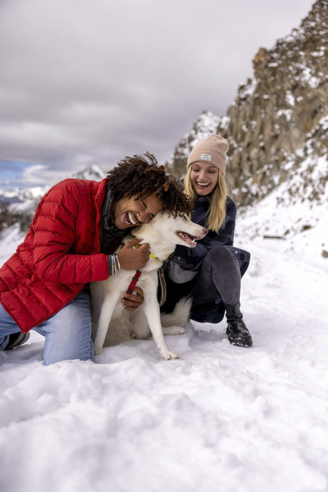 Ein fröhliches Paar spielt in einer verschneiten Bergregion mit einem glücklichen weißen Hund. Der Mann in einer roten Jacke kniet nieder und streichelt den Hund, während die Frau in einer dunklen Jacke und einer beigen Mütze lächelnd neben ihnen hockt. Im Hintergrund sind schneebedeckte Felsen und ein bewölkter Himmel zu sehen. © Fotografie Tomas Rodriguez