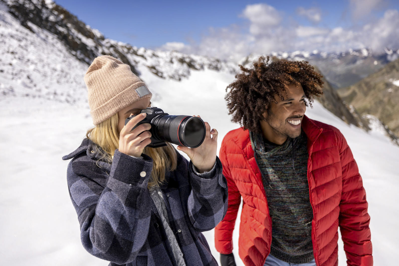 Zwei Menschen stehen auf einem schneebedeckten Berg. Einer trägt eine Beanie-Mütze und eine karierte Jacke und macht mit einer Kamera ein Foto. Der andere, in einer roten Jacke, steht neben ihnen, lächelt und blickt in die Ferne. Im Hintergrund sind schneebedeckte Gipfel und ein teilweise bewölkter blauer Himmel zu sehen. © Fotografie Tomas Rodriguez