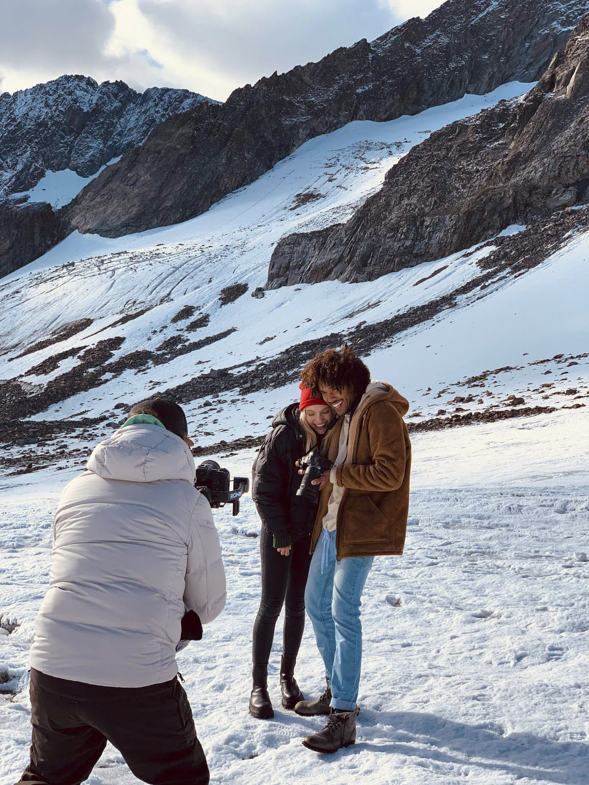 Ein Fotograf in einer weißen Jacke fotografiert ein lächelndes Paar in Winterkleidung, das eng beieinander in einer verschneiten Berglandschaft steht. Das Paar scheint den Moment vor der malerischen Kulisse aus schneebedeckten Gipfeln und zerklüftetem Gelände zu genießen. © Fotografie Tomas Rodriguez
