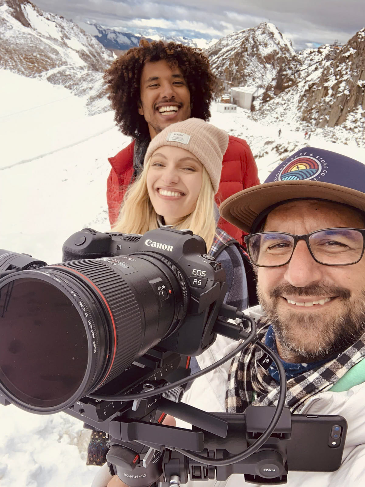 Drei Personen lächeln und posieren für ein Selfie auf einem schneebedeckten Berg. Die Person vorne hält eine Canon EOS R6-Kamera mit einem großen Objektiv. Die Person in der Mitte trägt eine beigefarbene Mütze und die Person hinten eine rote Jacke. Im Hintergrund sind schneebedeckte Gipfel zu sehen. © Fotografie Tomas Rodriguez