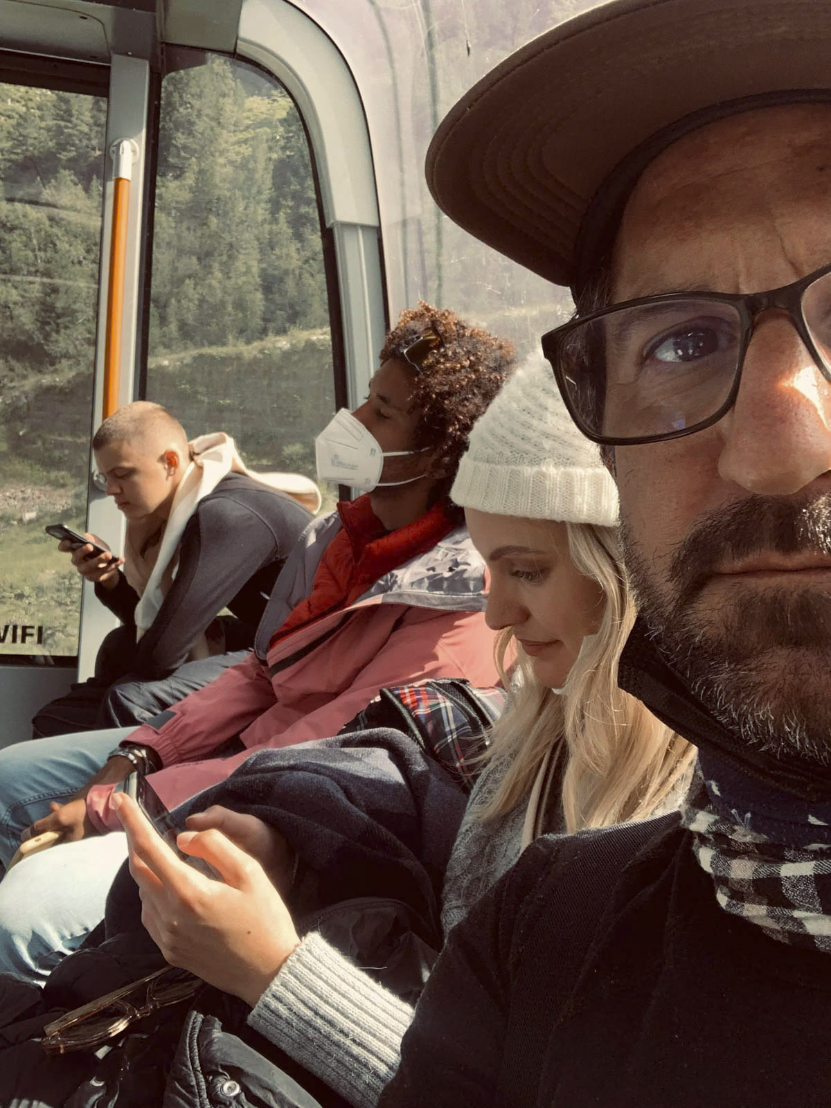 Eine Gruppe von vier Personen, drei Männer und eine Frau, sitzt in einem öffentlichen Verkehrsmittel, möglicherweise einem Bus oder Zug. Drei von ihnen sind auf ihre Telefone konzentriert, während ein Mann im Vordergrund mit Brille und Hut ein Selfie macht. © Fotografie Tomas Rodriguez