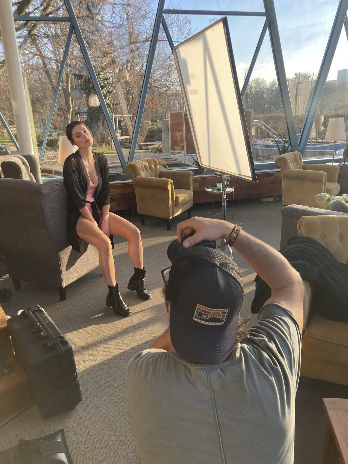 Ein Fotoshooting in einem Raum mit Glaswänden. Ein Model in einem schwarzen Bademantel und schwarzen Stiefeln sitzt auf einem Stuhl und posiert, während ein Fotograf mit Mütze ein Foto von ihr macht. Der Raum ist mit verschiedenen Stühlen und einer Leinwand im Hintergrund ausgestattet. Draußen sind Bäume zu sehen. © Fotografie Tomas Rodriguez
