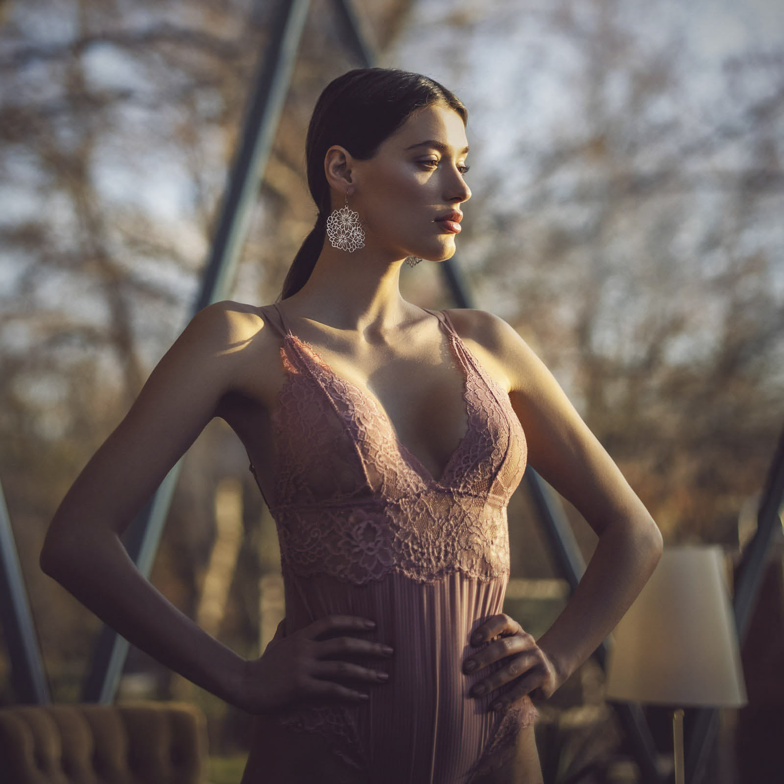 Eine Frau steht selbstbewusst da, die Hände in die Hüften gestemmt, trägt ein ärmelloses Spitzenkleid und große, kunstvolle Ohrringe. Sie ist in natürliches Licht getaucht, mit einem verschwommenen Hintergrund aus Bäumen und geometrischen Fensterstrukturen, die eine elegante und heitere Atmosphäre schaffen. © Fotografie Tomas Rodriguez