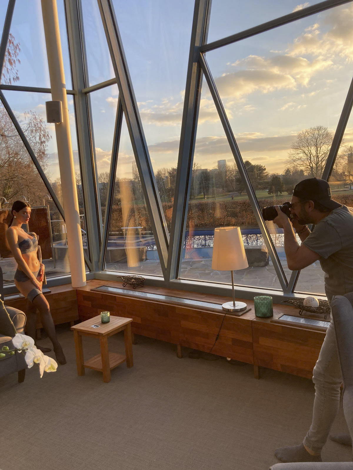 Ein Fotograf mit Mütze und grauem Hemd fotografiert eine Frau im Kleid. Sie befinden sich in einem Raum mit großen dreieckigen Fenstern, die einen malerischen Blick auf einen Park mit Bäumen und einem Teich bieten. Eine Holzbank, eine Lampe und ein kleiner Tisch mit einer Pflanze sind ebenfalls zu sehen. © Fotografie Tomas Rodriguez