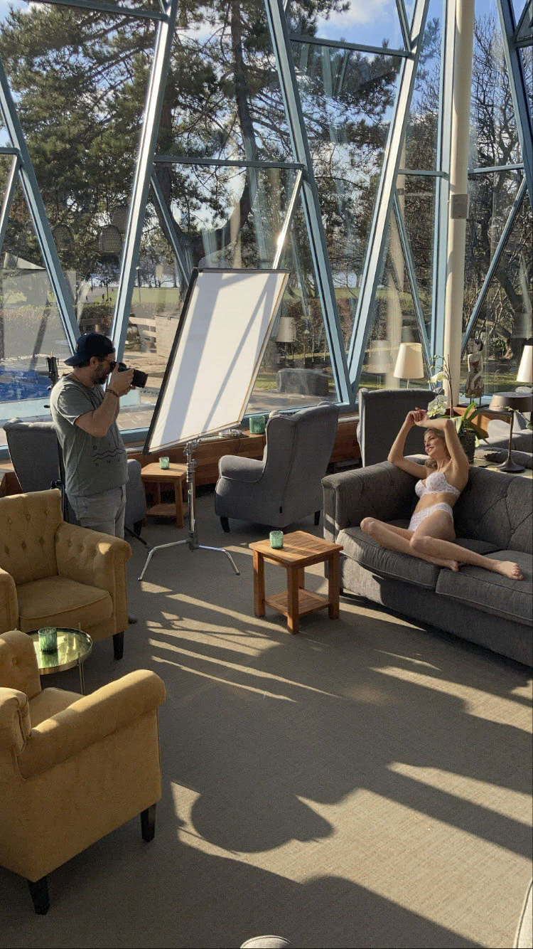 Ein Fotograf fotografiert ein Model, das auf einem grauen Sofa in einem Raum mit großen, dreieckigen Glasfenstern posiert. Das Model trägt weiße Dessous und hat die Arme erhoben. Die Kulisse besteht aus Sesseln, einem Beistelltisch und natürlichem Licht, das durch die Fenster hereinfällt. © Fotografie Tomas Rodriguez