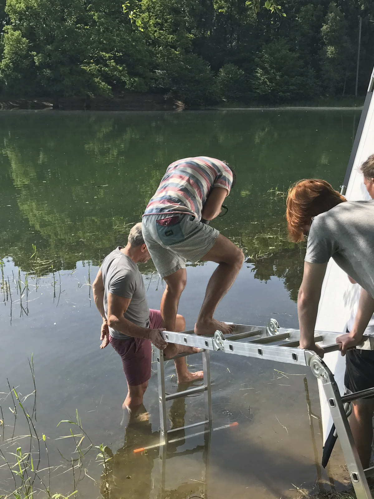 Drei Personen stehen am Ufer eines Sees: Ein Mann steht im seichten Wasser und hält eine Leiter, während ein anderer Mann die Leiter hochklettert und eine dritte Person die Leiter vom Ufer aus stützt. Im Hintergrund sind Bäume und Grün zu sehen, und das Wasser ist ruhig. © Fotografie Tomas Rodriguez