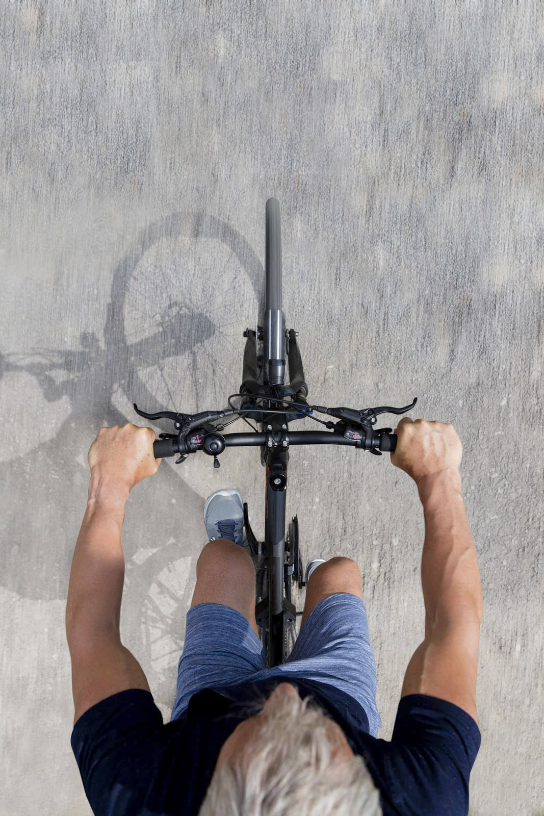 Eine Draufsicht auf eine Person mit grauem Haar, die auf einem gepflasterten Weg Fahrrad fährt. Die Person umklammert mit den Händen den Lenker und trägt ein blaues Hemd und blaue Shorts. Das Fahrrad wirft einen Schatten auf den Boden. © Fotografie Tomas Rodriguez
