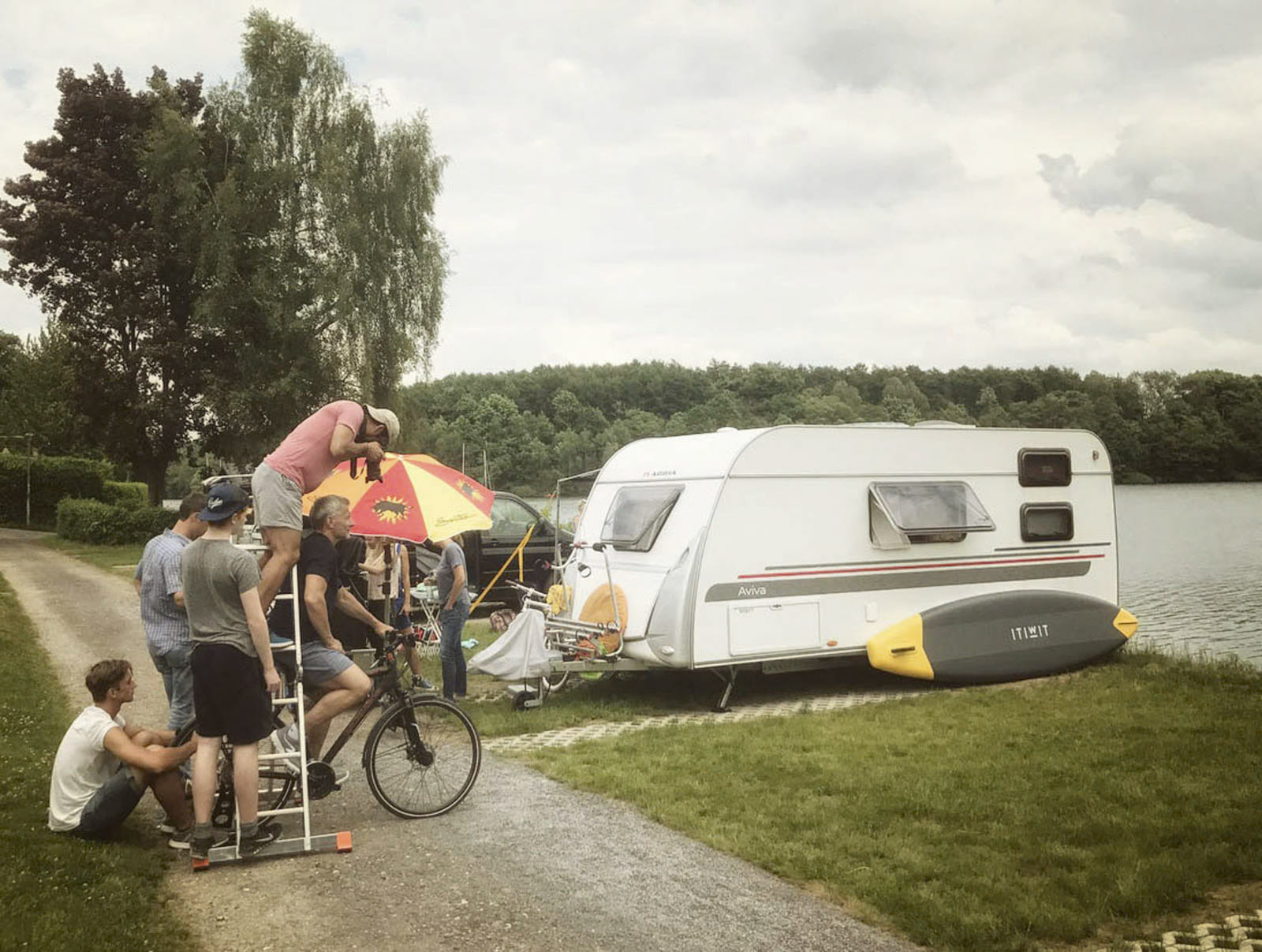 Eine Gruppe von Menschen versammelt sich vor einem stillstehenden weißen Wohnwagen in der Nähe eines Sees. Eine Person richtet eine Kamera auf einem Stativ auf einer Trittleiter aus. Andere sind in der Nähe, einige sitzen und einige stehen. Neben dem Wohnwagen ist ein Paddleboard zu sehen. © Fotografie Tomas Rodriguez