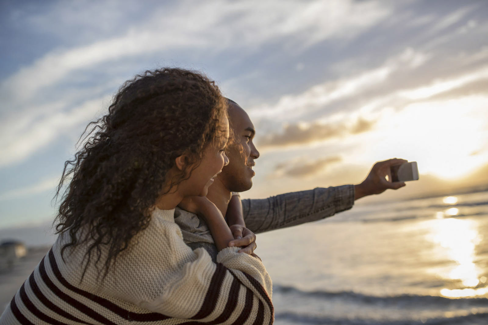 Ein Paar steht bei Sonnenuntergang an einem Strand. Eine Person trägt die andere huckepack, während beide lächeln und ein Selfie mit einem Smartphone machen. Im Hintergrund sind das Meer und ein bunter Himmel mit Wolken zu sehen. © Fotografie Tomas Rodriguez