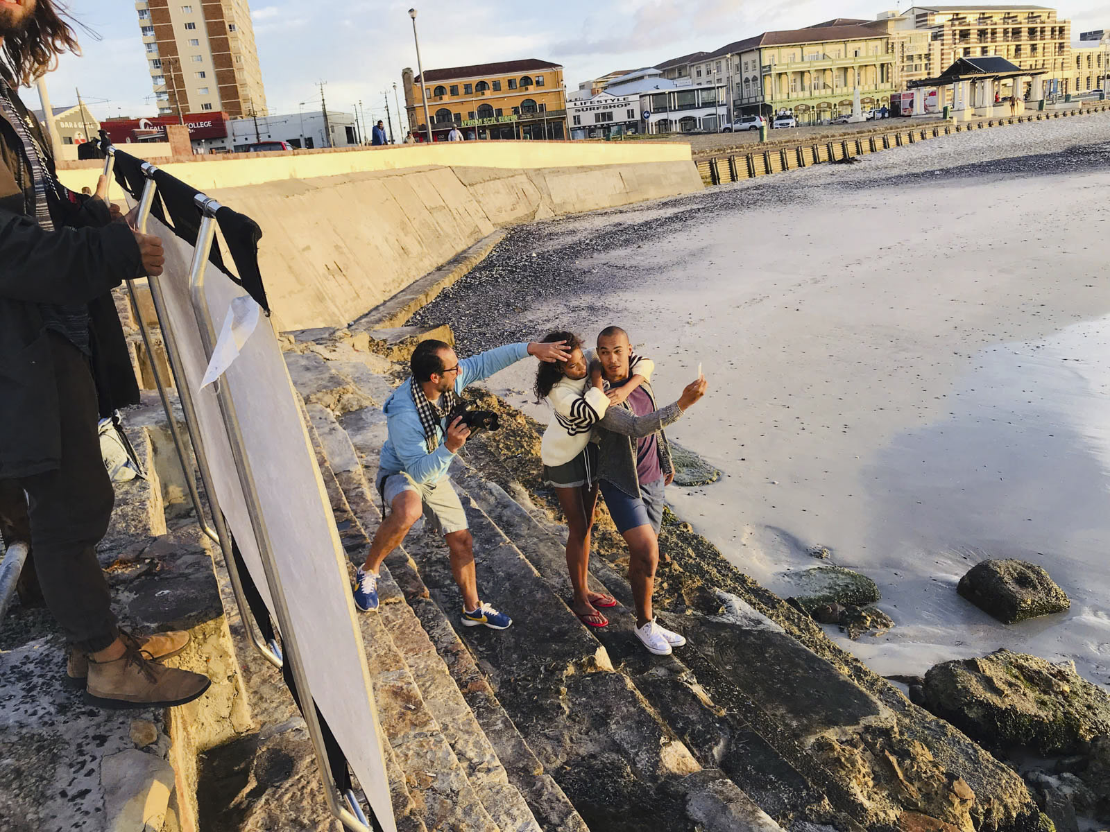 Drei Personen posieren energisch bei Sonnenuntergang an einem felsigen Strand. Eine Person hält eine Kamera und eine andere Person macht dynamische Posen. Die dritte Person steht neben einem reflektierenden Diffusor, den eine andere Person hält. Im Hintergrund sind Gebäude und das Meer zu sehen. © Fotografie Tomas Rodriguez
