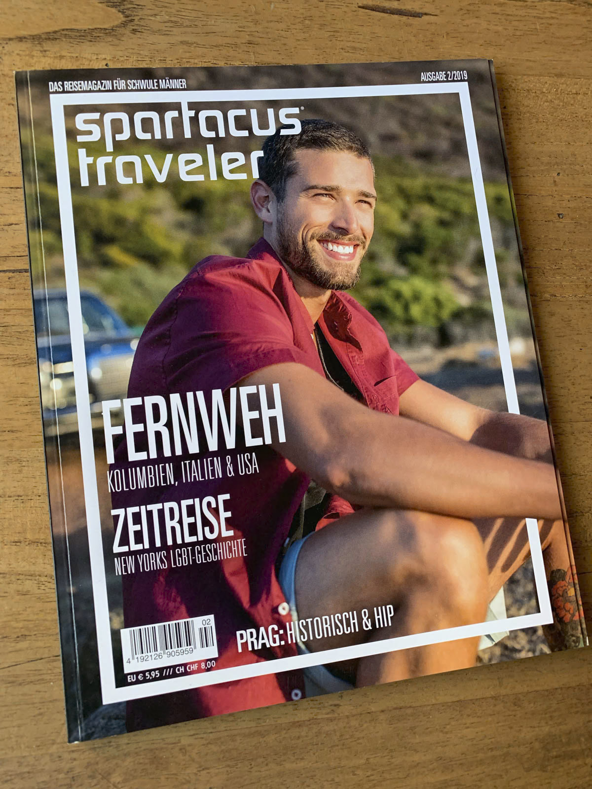 Auf dem Cover einer Zeitschrift mit dem Titel „Spartacus Traveler“ ist ein lächelnder Mann zu sehen, der im Freien sitzt. Im Text werden Reiseziele wie Kolumbien, Italien und die USA erwähnt, und ein Schwerpunkt liegt auf der LGBT-Geschichte New Yorks. Die Ausgabe stammt aus dem Jahr 2018. © Fotografie Tomas Rodriguez