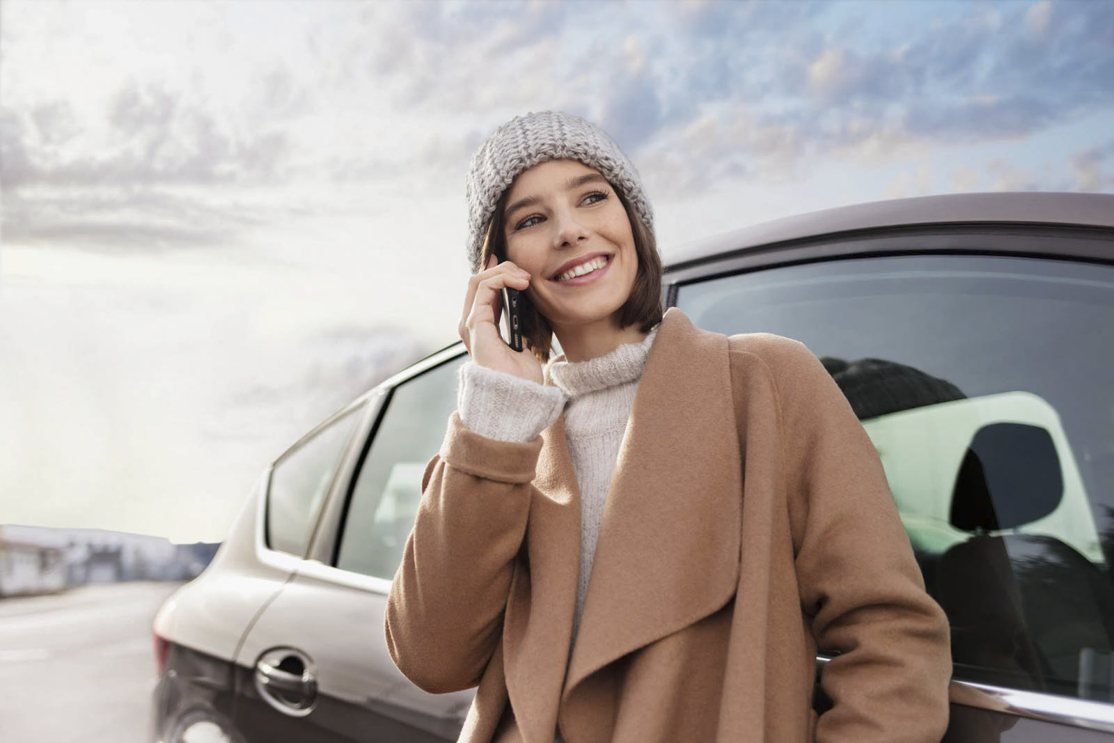 Eine lächelnde Frau in einem beigen Mantel und einer Strickmütze steht draußen neben einem Auto und telefoniert. Der Himmel ist bewölkt mit leichten blauen Schattierungen. Sie wirkt glücklich und ist in das Gespräch vertieft. © Fotografie Tomas Rodriguez