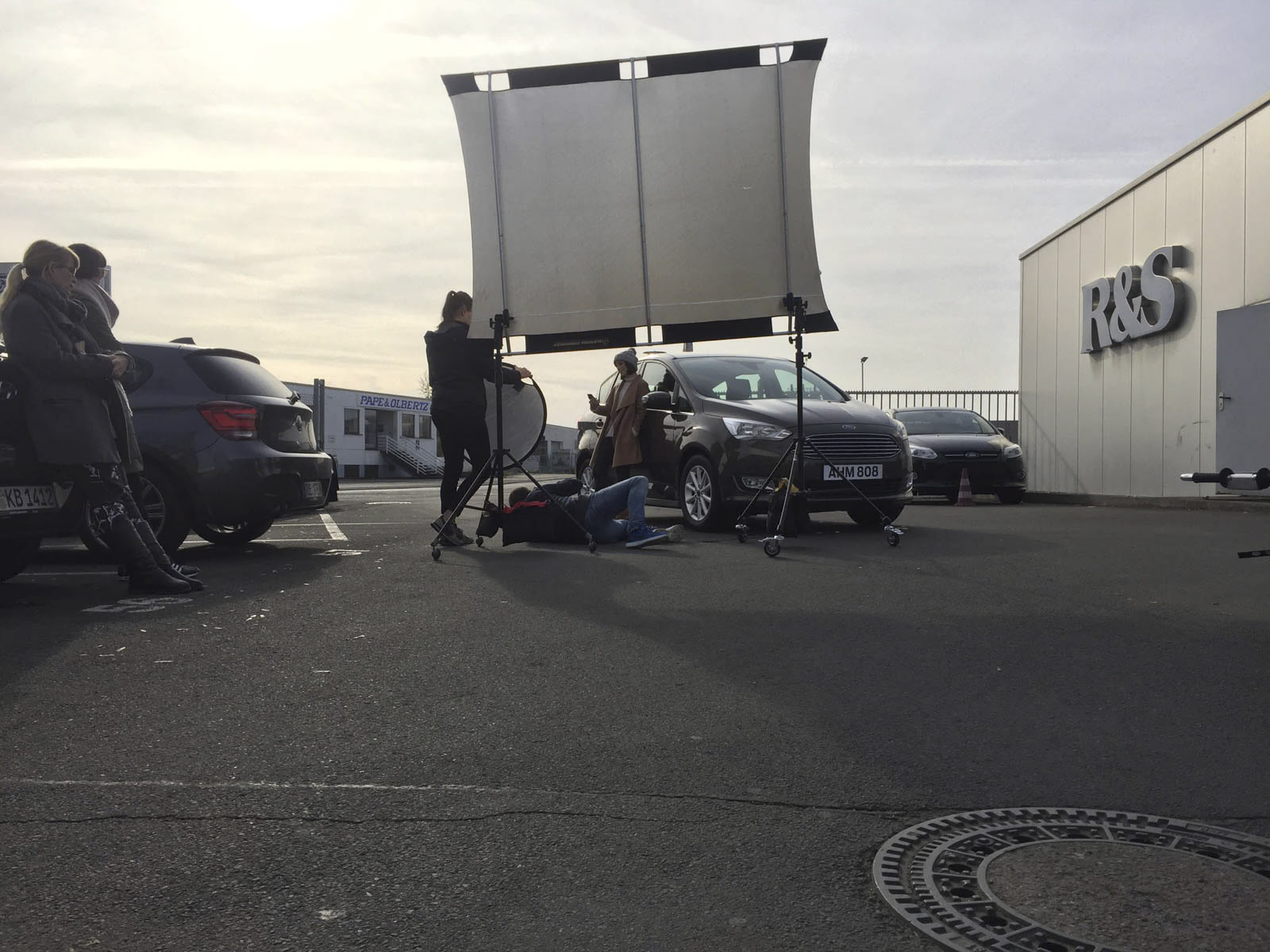 Einige Leute haben sich im Freien um ein Auto versammelt, eine Person liegt auf dem Boden unter dem Auto. In der Nähe ist eine große Reflektortafel aufgestellt, um die Beleuchtung anzupassen. Der Ort scheint ein Parkplatz neben einem Gebäude mit der Aufschrift „R&S“ zu sein. © Fotografie Tomas Rodriguez