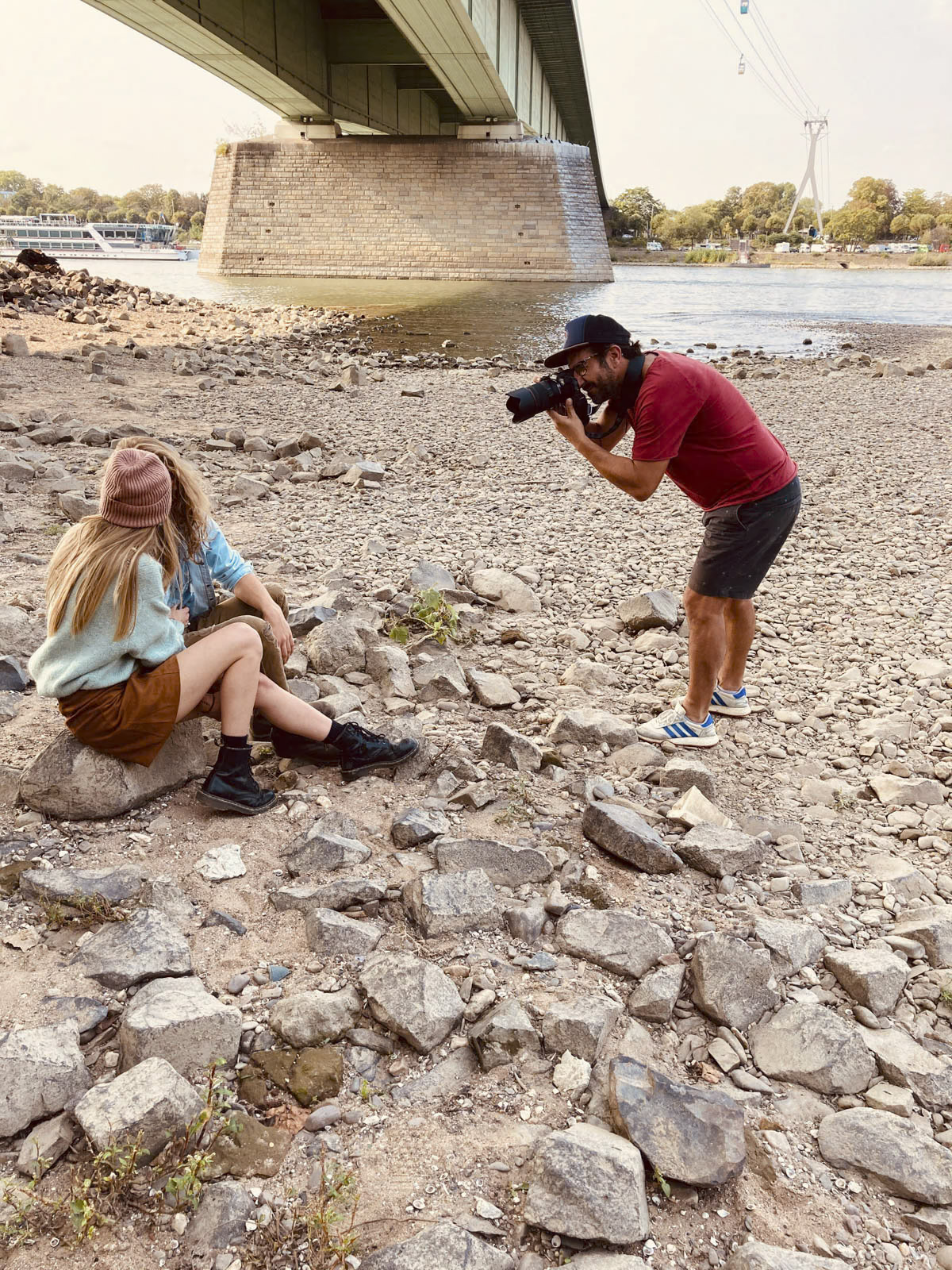 Ein Fotograf in rotem Hemd und Shorts fotografiert zwei Personen, die an einem felsigen Flussufer sitzen. Sie befinden sich unter einer großen Brücke mit Bäumen und Wasser im Hintergrund. Eine Person trägt eine rosa Beanie und braune Shorts, während die andere einen Hut und ein blaues Hemd trägt. © Fotografie Tomas Rodriguez