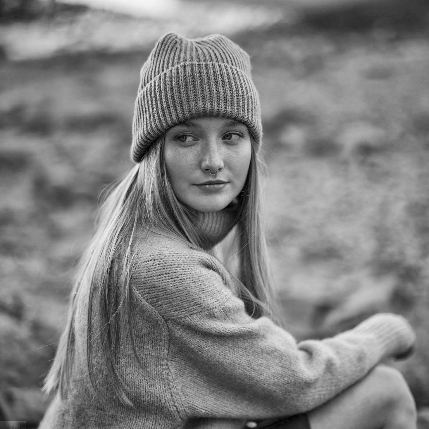 Eine Frau mit langen Haaren, die eine Strickmütze und einen kuscheligen Pullover trägt, blickt nach links. Der Hintergrund ist verschwommen und scheint eine Außenkulisse mit undeutlichen Naturelementen zu sein. Das Bild ist schwarz-weiß, was ihm ein zeitloses und besinnliches Gefühl verleiht. © Fotografie Tomas Rodriguez