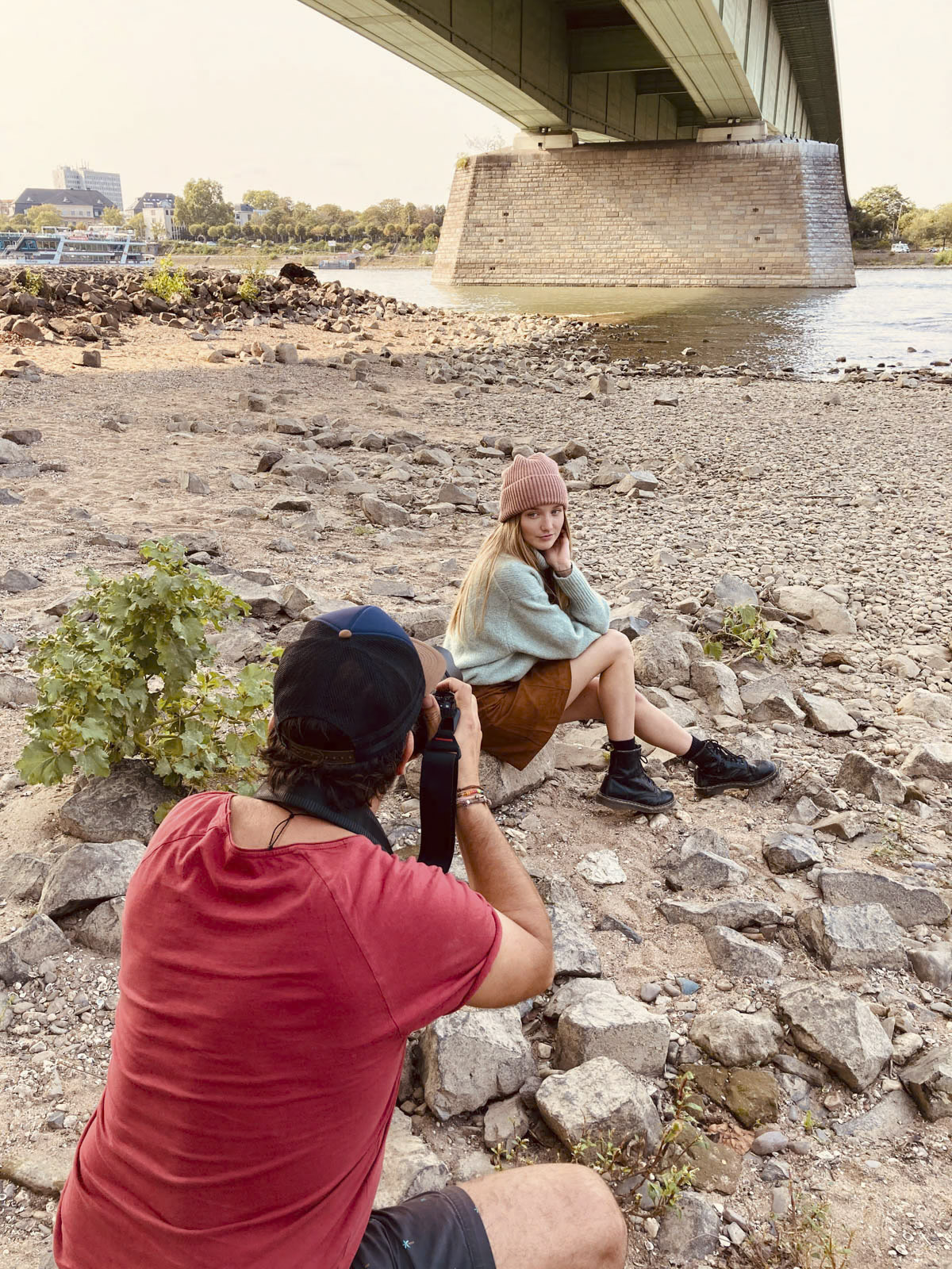 Ein Fotograf in rotem Hemd und schwarzem Hut fotografiert eine Frau, die auf Felsen am Flussufer sitzt. Die Frau trägt eine Beanie-Mütze und blickt in die Kamera. Im Hintergrund überspannt eine große Brücke den Fluss, und das Ufer ist mit verstreuten Steinen bedeckt. © Fotografie Tomas Rodriguez