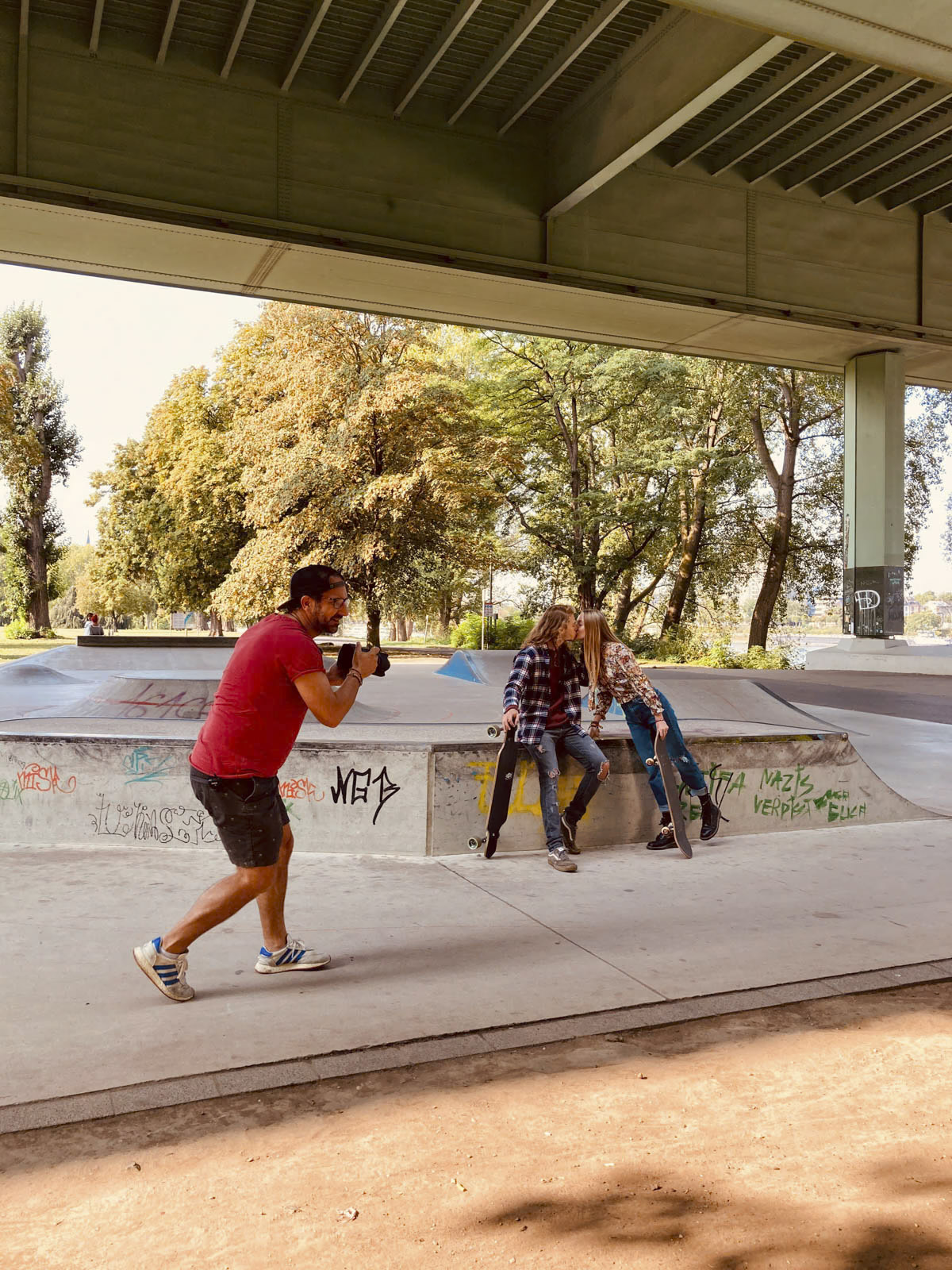 Eine Person in einem roten Hemd fotografiert zwei Personen, die am Rand einer Skaterampe unter einer Brücke sitzen. Im Hintergrund sind Bäume und mit Graffiti bedeckte Betonstrukturen zu sehen. © Fotografie Tomas Rodriguez