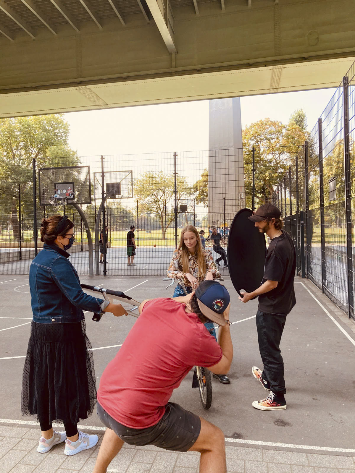 Menschen haben sich auf einem überdachten Basketballplatz im Freien versammelt. Im Mittelpunkt steht ein Mädchen auf einem Fahrrad. Um sie herum halten andere Leute verschiedene Filmausrüstungen. Im Hintergrund sind hinter dem Zaun Bäume und ein Teil eines Gebäudes zu sehen. © Fotografie Tomas Rodriguez