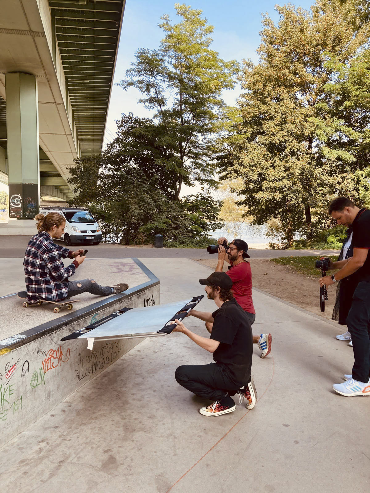 Ein Skateboarder sitzt auf einer Rampe unter einer Überführung, während sich drei Personen mit Kameras um ihn herum versammeln, um zu filmen oder zu fotografieren. Eine Person kniet mit einer Kamera, eine andere hockt mit ihrer Ausrüstung und die dritte steht da und fängt die Szene ein. Im Hintergrund sind Bäume und ein geparktes Auto zu sehen. © Fotografie Tomas Rodriguez