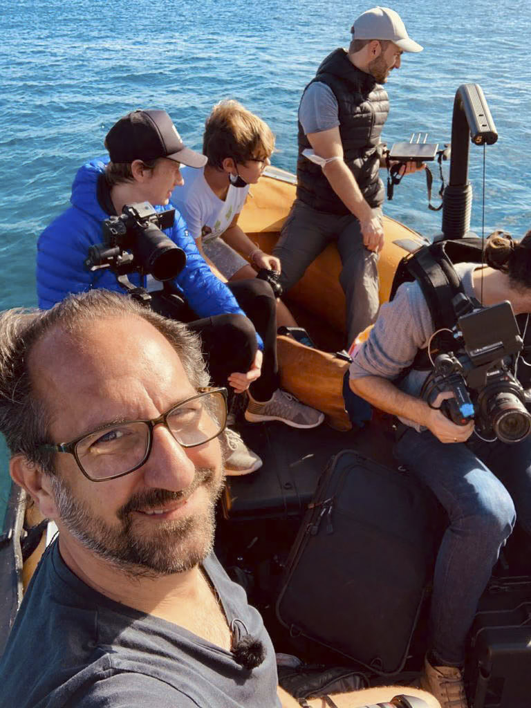 Eine Gruppe von vier Männern ist mit Kameras und Filmausrüstung auf einem Boot. Ein Mann macht ein Selfie, während die anderen damit beschäftigt sind, ihre Kameraausrüstung einzustellen und zu positionieren. Die Szene spielt vor dem Hintergrund ruhigen blauen Wassers unter einem klaren Himmel. © Fotografie Tomas Rodriguez