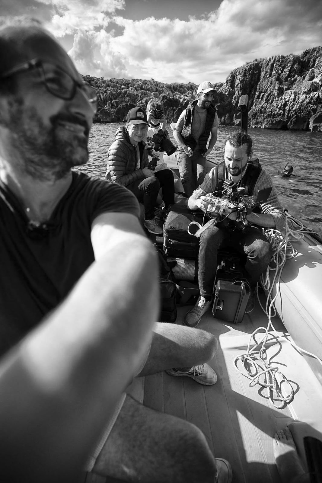 Schwarzweißbild einer Gruppe von Menschen auf einem kleinen Boot mit Ausrüstung. Eine Person macht ein Selfie und zeigt andere hinter ihr, die mit unterschiedlicher Ausrüstung und Seilen arbeiten. Sie sind von Wasser und felsigen Klippen unter einem teilweise bewölkten Himmel umgeben. © Fotografie Tomas Rodriguez