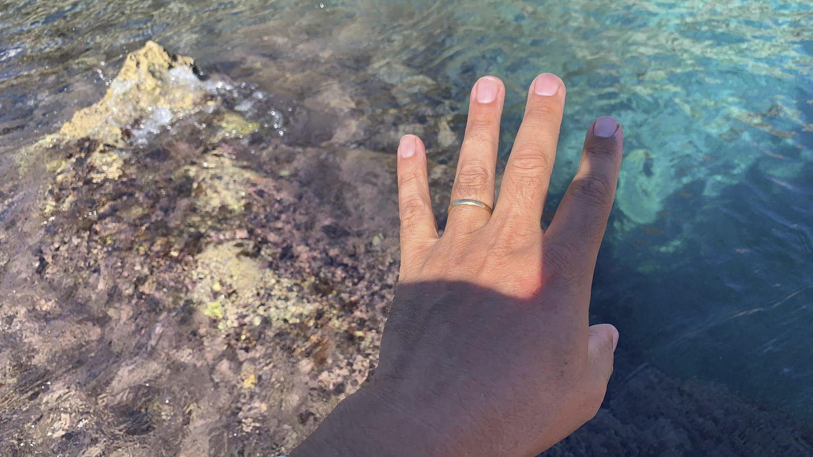 Eine Hand mit einem einfachen, dünnen Ring am Ringfinger ist über klares, seichtes Wasser ausgestreckt, unter dem sich eine felsige, algenbedeckte Oberfläche befindet. Das Sonnenlicht scheint hell auf die Hand und das Wasser und hebt den klaren blauen Farbton des Wassers hervor. © Fotografie Tomas Rodriguez