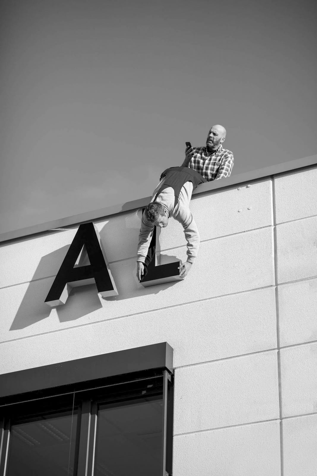 Ein Mann im karierten Hemd lehnt sich über die Kante eines Gebäudes und hält einen anderen Mann am Arm. Der zweite Mann hängt und greift nach dem Buchstaben „L“ neben dem Buchstaben „A“ an der Fassade des Gebäudes. Das Bild ist schwarzweiß. © Fotografie Tomas Rodriguez