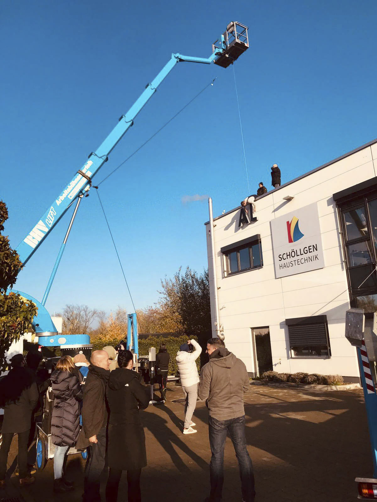Eine Gruppe von Menschen fotografiert und beobachtet einen Mann, der mit einer blauen Hubarbeitsbühne auf das Dach eines Gebäudes mit der Aufschrift „Schöllgen Haustechnik“ gehoben wird. Eine weitere Person steht am Rand des Dachs. Das Wetter scheint sonnig und klar. © Fotografie Tomas Rodriguez