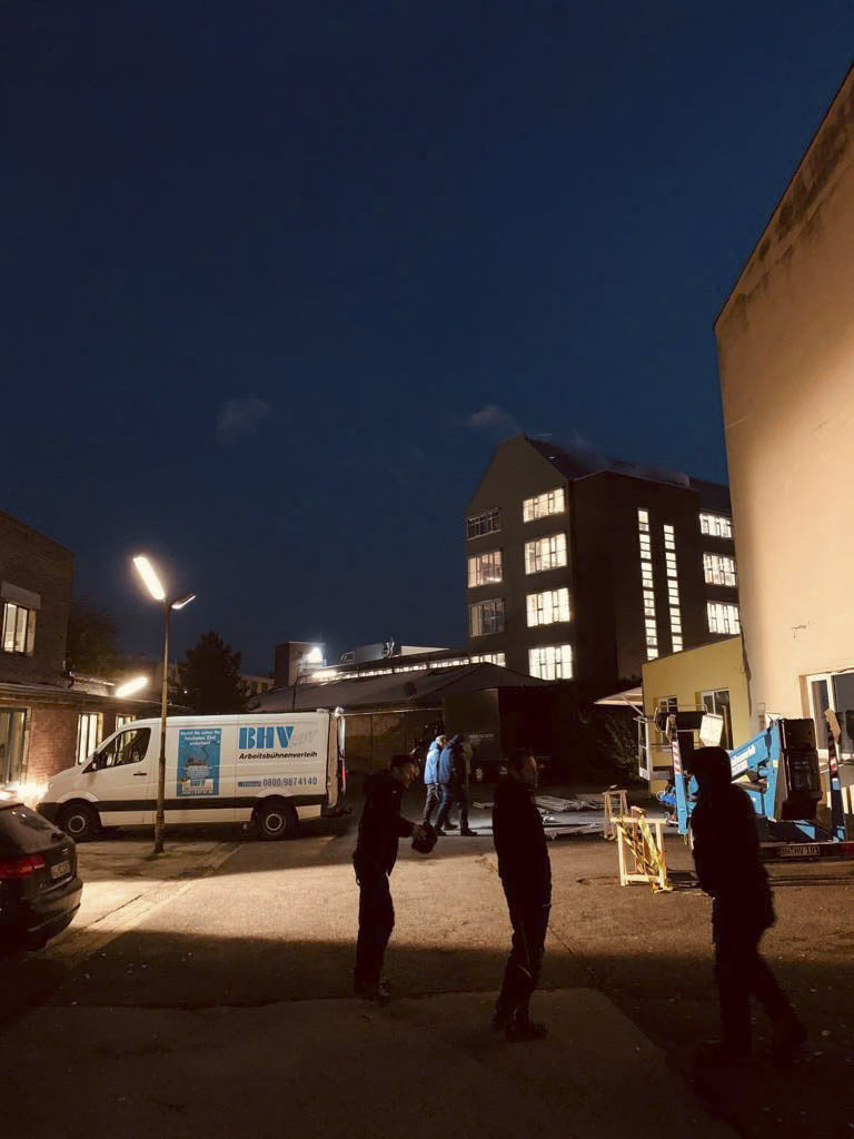 Eine Nachtszene, die eine gut beleuchtete Gasse mit Schatten von drei stehenden Personen zeigt. Ein weißer Lieferwagen mit blauen Markierungen ist auf der linken Seite neben einem Gebäude geparkt. Im Hintergrund sind hohe Gebäude mit beleuchteten Fenstern vor einem dunklen Himmel zu sehen. © Fotografie Tomas Rodriguez