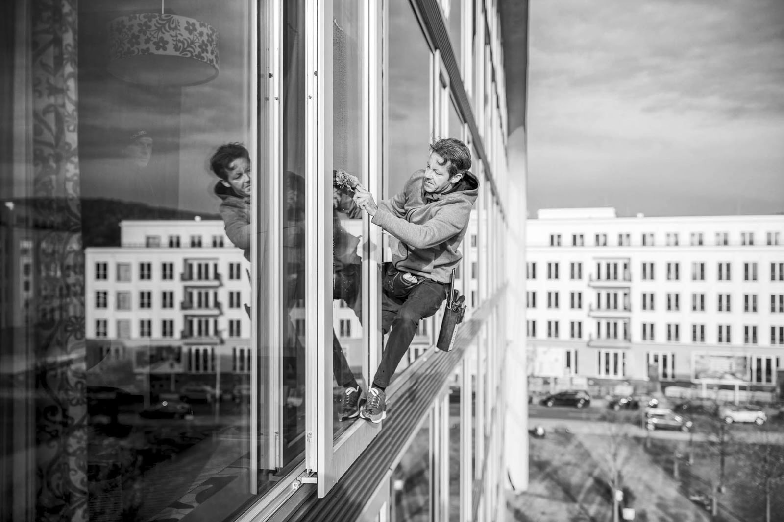 Eine Person hängt an einem Gurt vor einem hohen Gebäude und putzt die Fenster. Sie trägt einen Kapuzenpullover und Jeans und ist auf ihre Aufgabe konzentriert. Im Hintergrund sind mehrere mehrstöckige Gebäude und ein klarer Himmel zu sehen. Das Bild ist schwarzweiß. © Fotografie Tomas Rodriguez