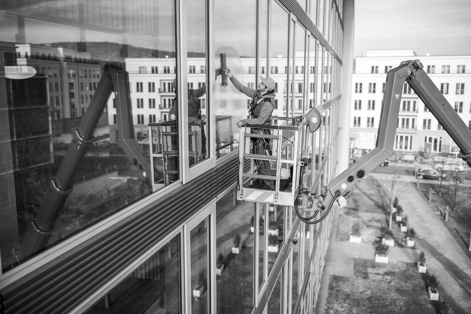 Ein Arbeiter mit Sicherheitsgurt und Helm putzt die Fenster eines hohen Gebäudes mit einem Abzieher, während er auf einer Hebebühne steht. Im Hintergrund sind mehrere moderne Gebäude zu sehen. © Fotografie Tomas Rodriguez