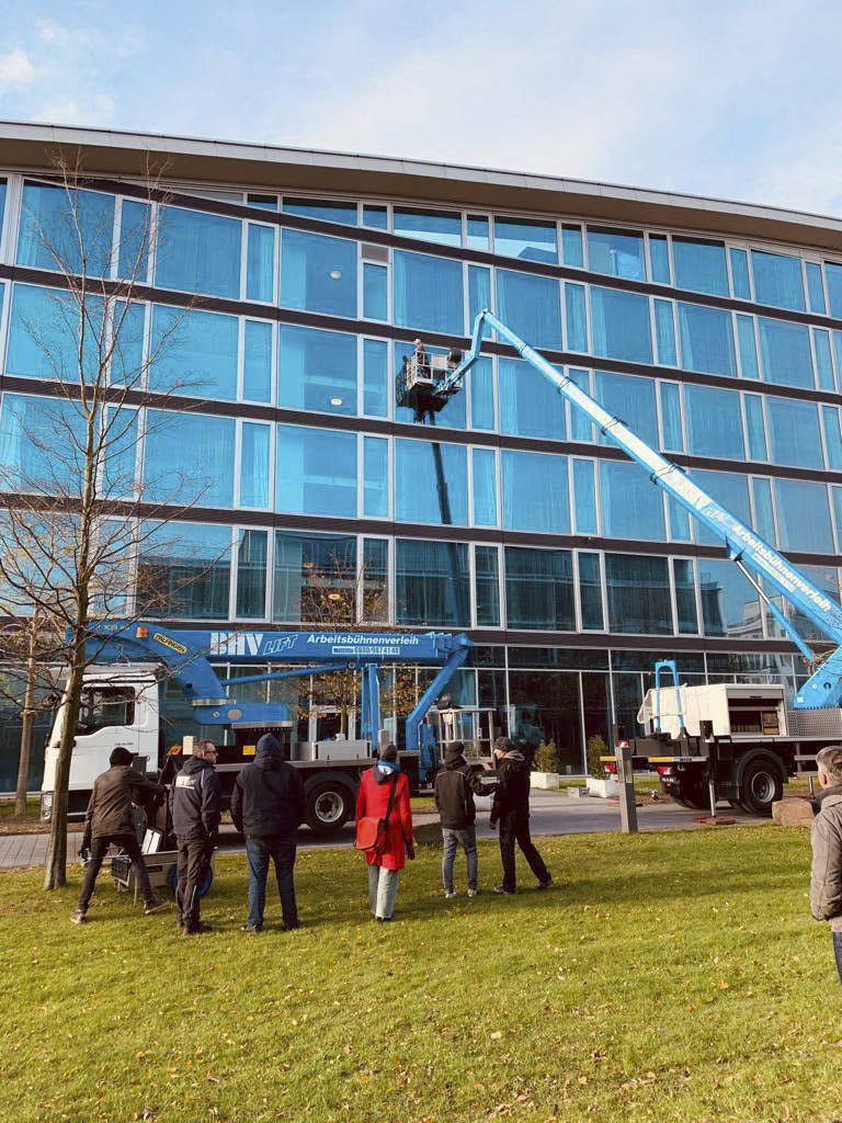 Arbeiter werden auf blauen hydraulischen Hebebühnen vor ein modernes Glasgebäude gehoben. Mehrere Leute am Boden beobachten die Arbeit, an klaren Tagen tragen einige von ihnen Mäntel. Auf der Wiese vor dem Gebäude steht auch ein Lastwagen mit Kran. © Fotografie Tomas Rodriguez