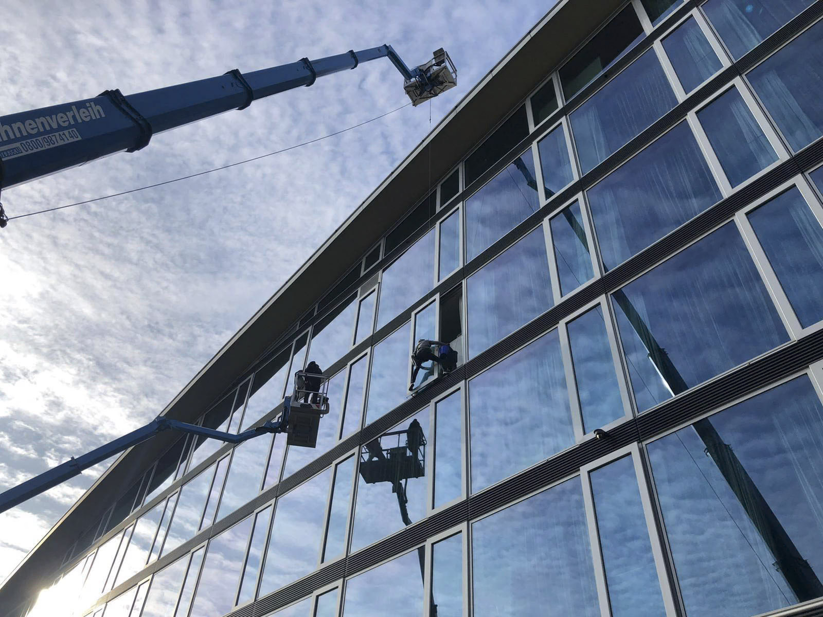 Drei Arbeiter auf Hebebühnen reinigen die Fenster eines hohen Gebäudes. Die Plattformen werden von blauen hydraulischen Aufzügen ausgefahren. Das Gebäude verfügt über große Glasscheiben, in denen sich ein teilweise bewölkter Himmel spiegelt. Die Szene spielt bei Tageslicht. © Fotografie Tomas Rodriguez