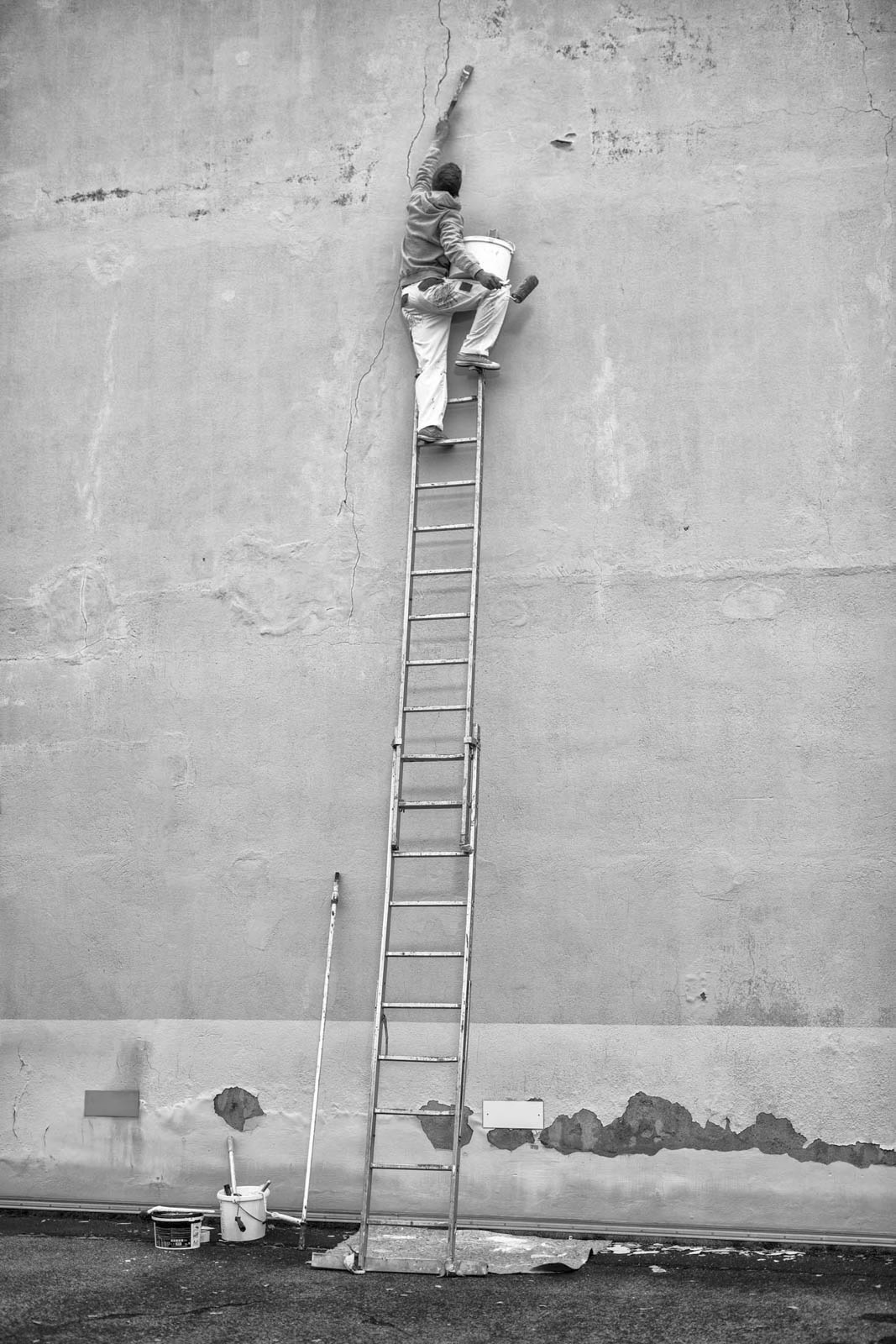 Eine Person in Arbeitskleidung klettert auf eine hohe Leiter, die an einer rissigen Wand lehnt. Sie greift mit einem Eimer in der Hand nach dem Riss. Am Fuß der Leiter stehen verschiedene Werkzeuge und ein kleines Gerüst. Das Bild ist schwarzweiß. © Fotografie Tomas Rodriguez