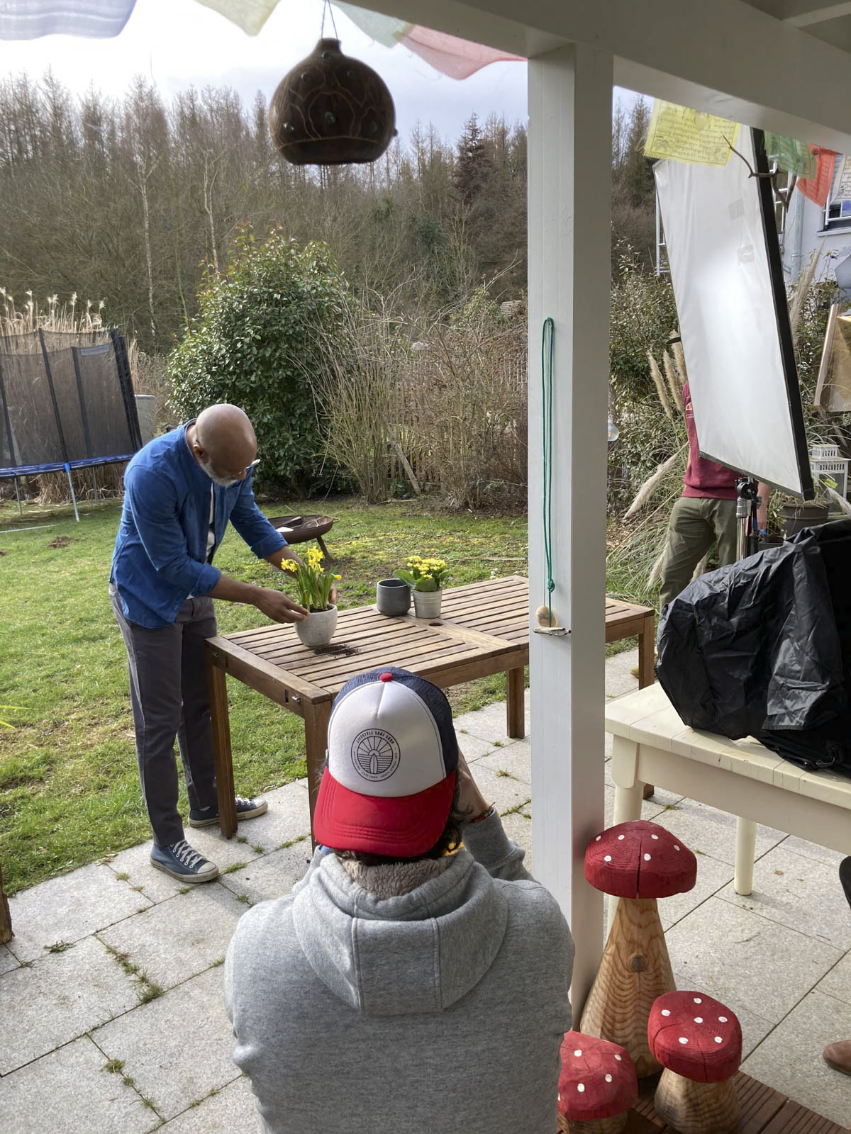 Eine Person in blauer Kleidung arrangiert draußen auf einem Holztisch einen Strauß gelber Blumen, während eine andere Person mit einer Baseballkappe ein Foto davon macht. Die Szene spielt in einem Garten mit Bäumen im Hintergrund und verschiedenen Gartengeräten in der Nähe. © Fotografie Tomas Rodriguez