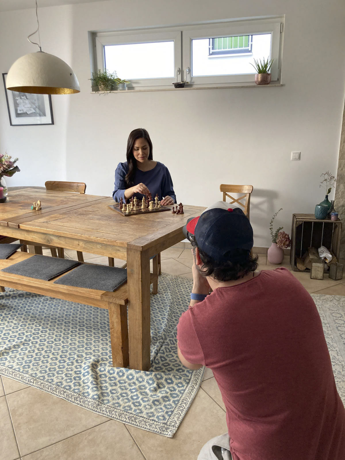 Eine Frau sitzt an einem Holztisch und spielt Schach, während ein Mann in rotem Hemd und Baseballkappe ein Foto von ihr macht. Der Raum ist durch ein Fenster gut beleuchtet. Zur Dekoration gehören Pflanzen, ein Teppich und verschiedene Möbel. © Fotografie Tomas Rodriguez